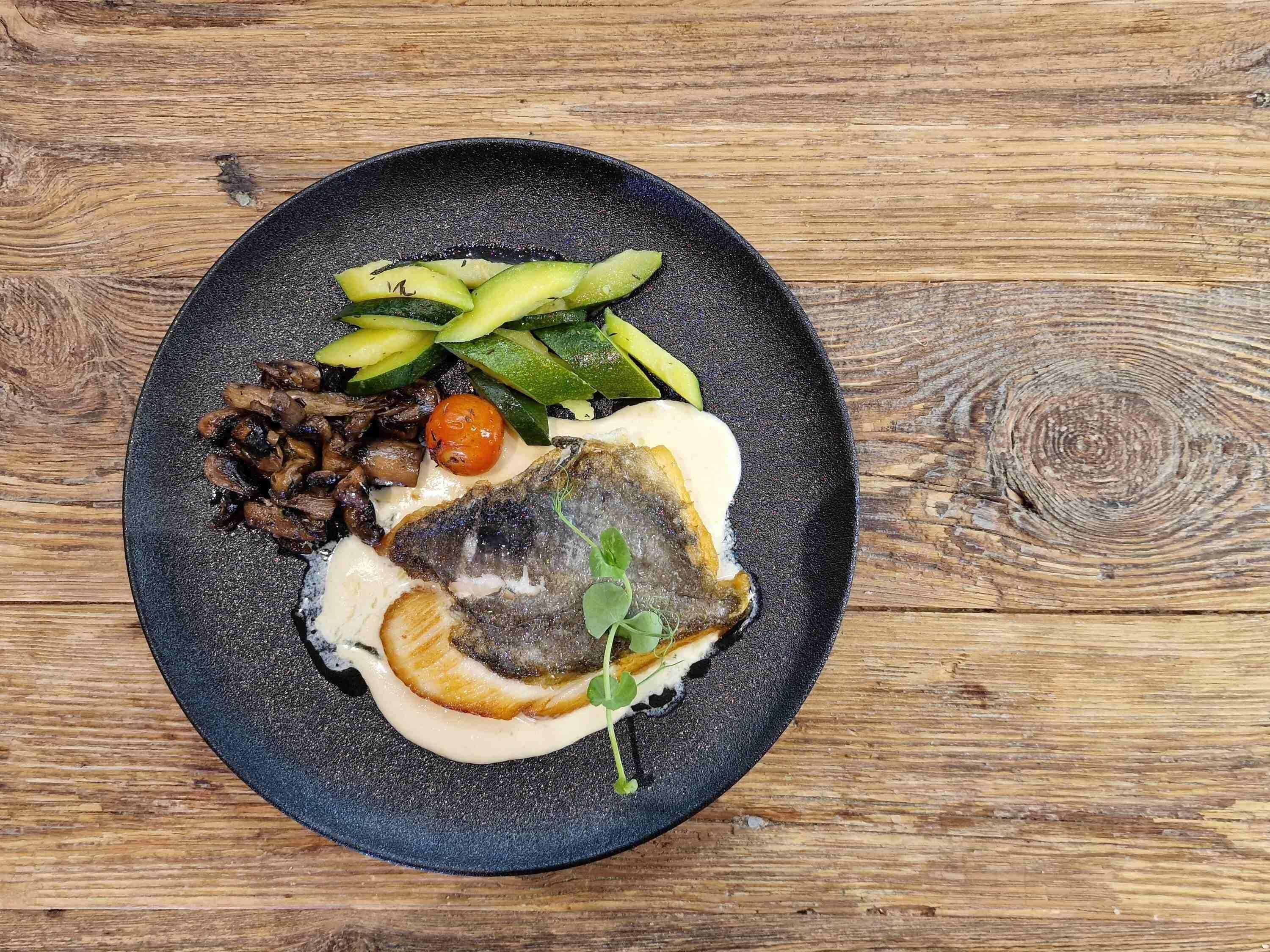 A plate with a fish, vegetables, and a small tomato on a wooden table.