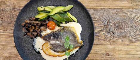 A plate with a fish, vegetables, and a small tomato on a wooden table.