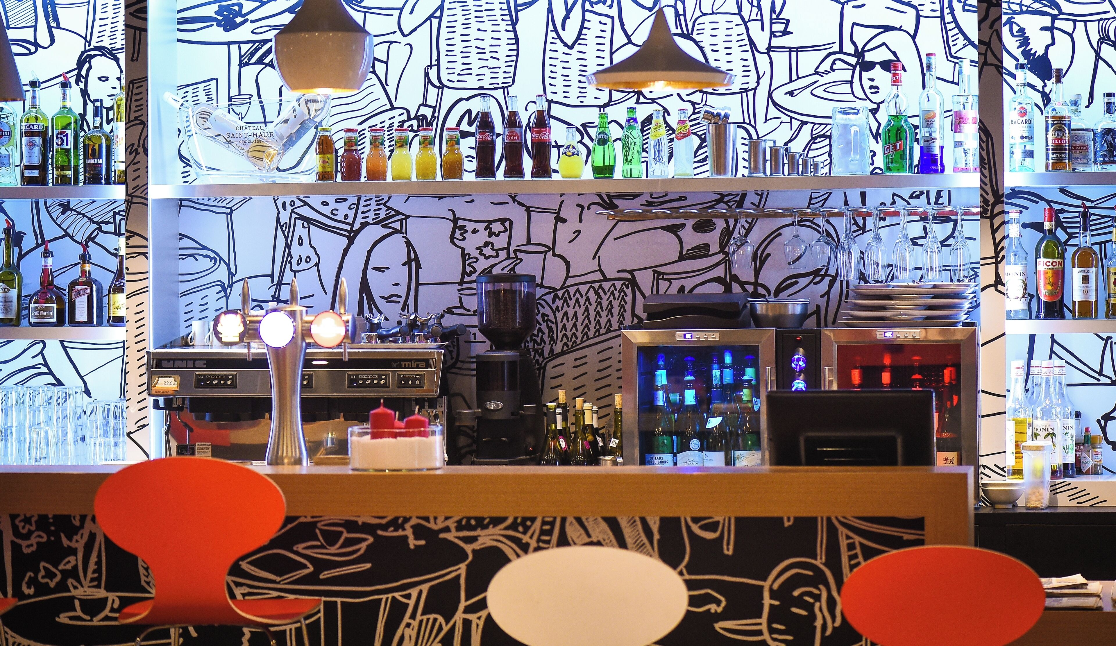Zona de bar con diferentes botellas y vasos, una máquina de café y una pared decorada con arte abstracto.