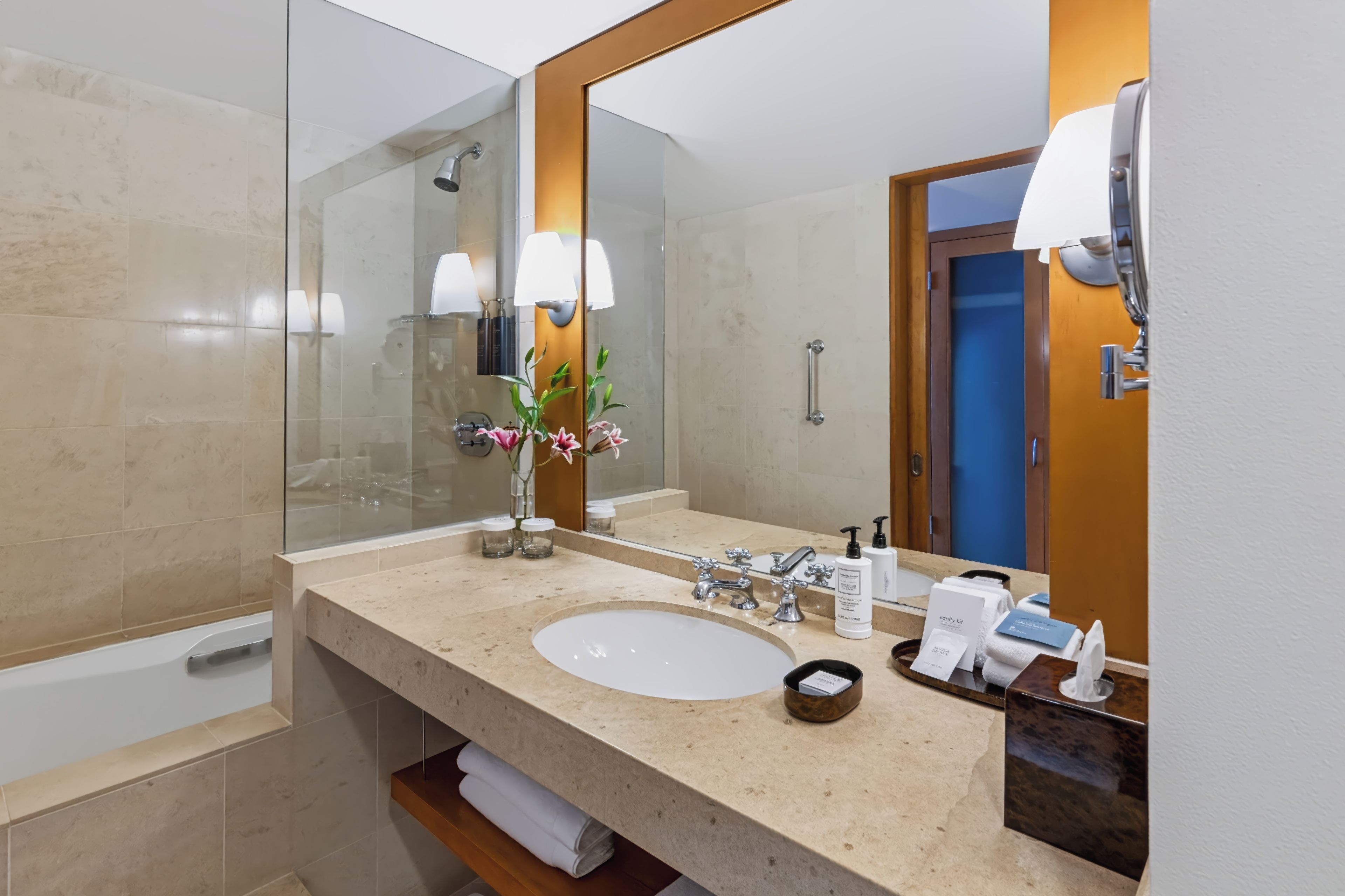Un baño de hotel con un espejo grande, un lavabo, una ducha y un tocador con artículos de aseo.