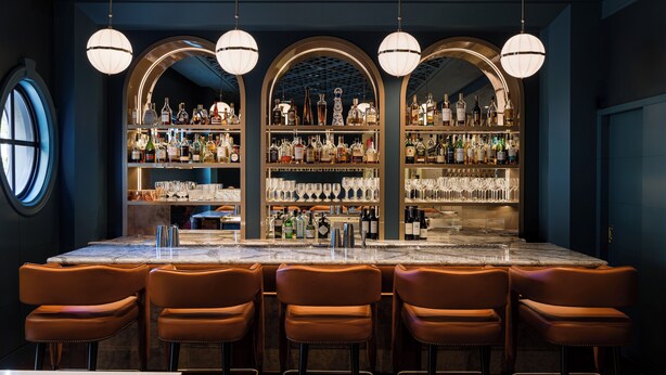 Una zona de bar con mostrador de mármol, taburetes de cuero y estantes llenos de botellas y copas.