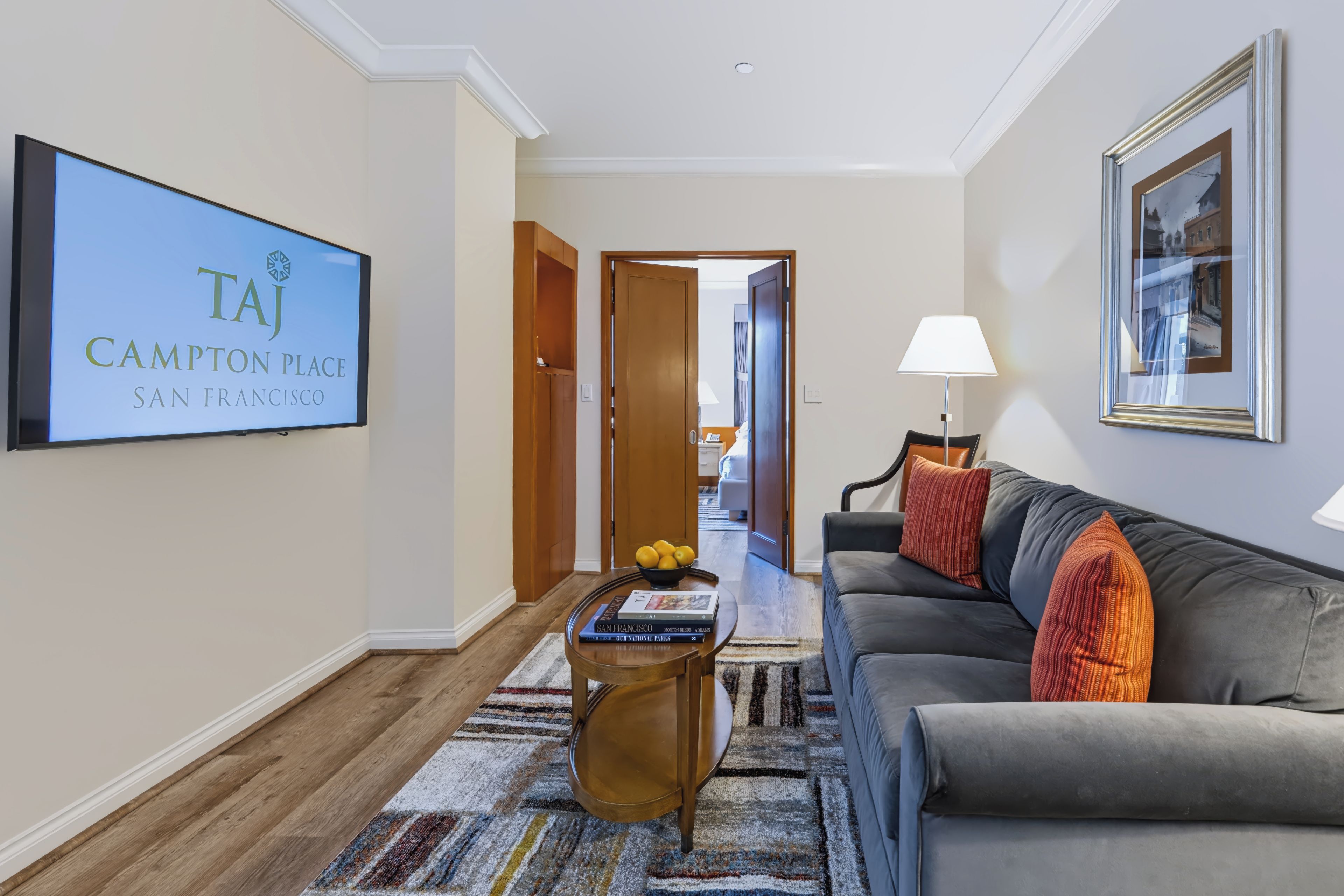 A hotel lobby with a flat-screen TV displaying 'TAJ CAMPTON PLACE SAN FRANCISCO', a sofa with a lamp, a coffee table with books, and a patterned rug.