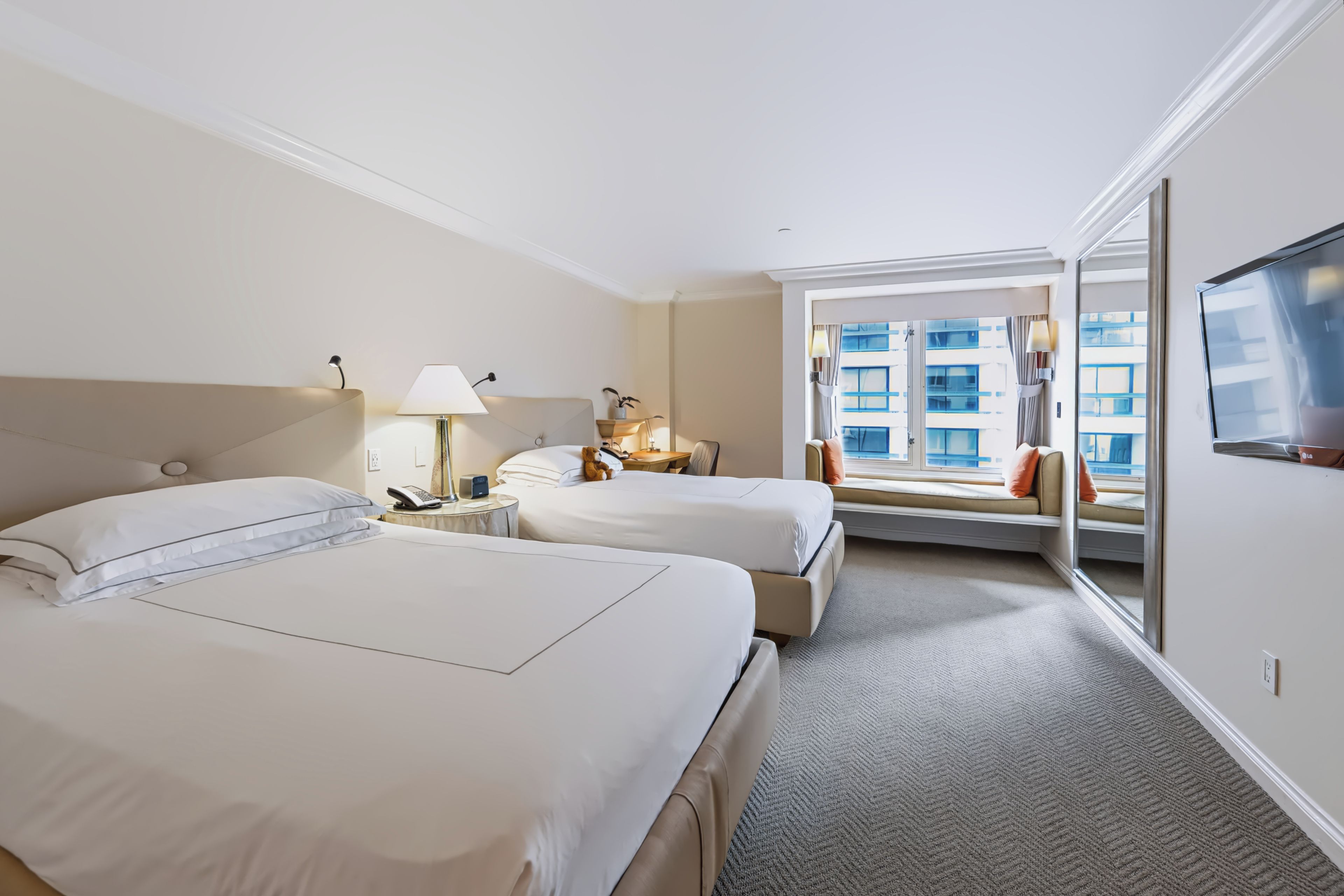 Habitación de hotel con dos camas, una mesita de noche con lámpara, ventana con vistas a edificios y televisión en la pared.