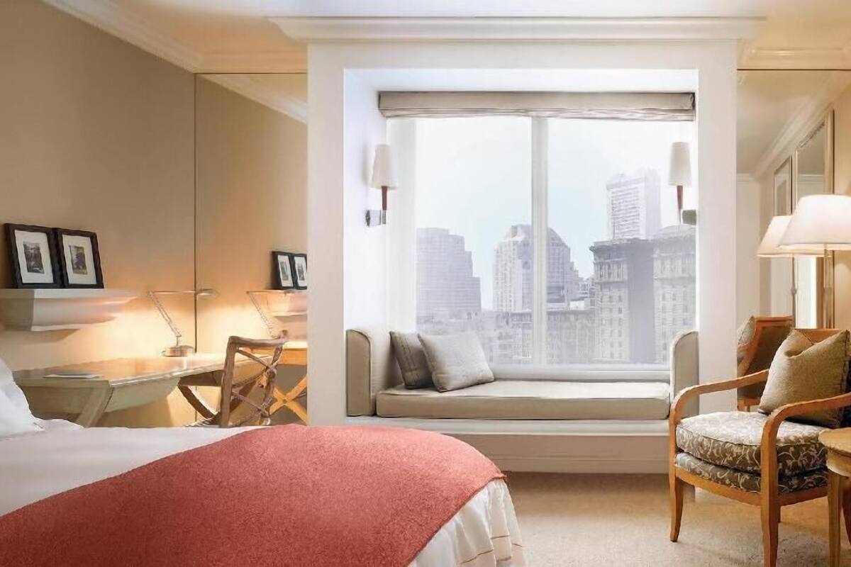 A hotel room with a bed, a desk, a chair, a window with a city view, and a sofa with a window seat.