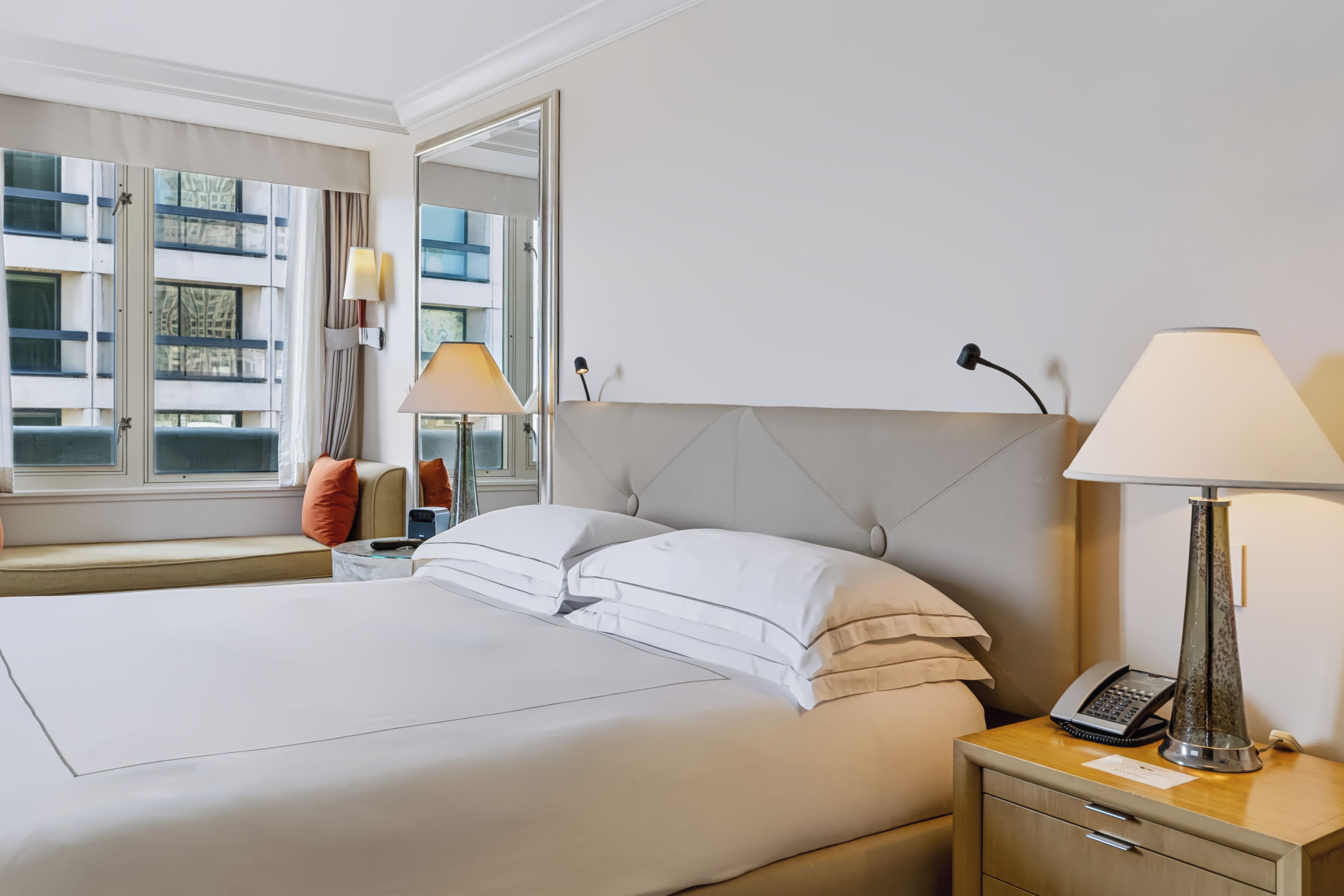 A hotel room with a large bed, bedside lamps, a nightstand with a phone, and a window with a view of buildings.