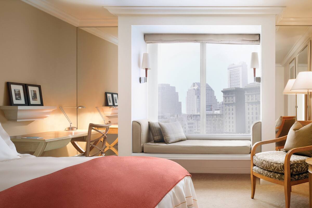 A hotel room with a bed, a desk with a lamp, a chair, a window with a city view, and a sofa with cushions.