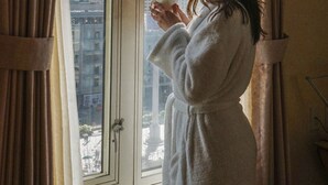 Una persona con bata blanca de pie junto a una ventana con cortinas, mirando un paisaje urbano con edificios y nieve.