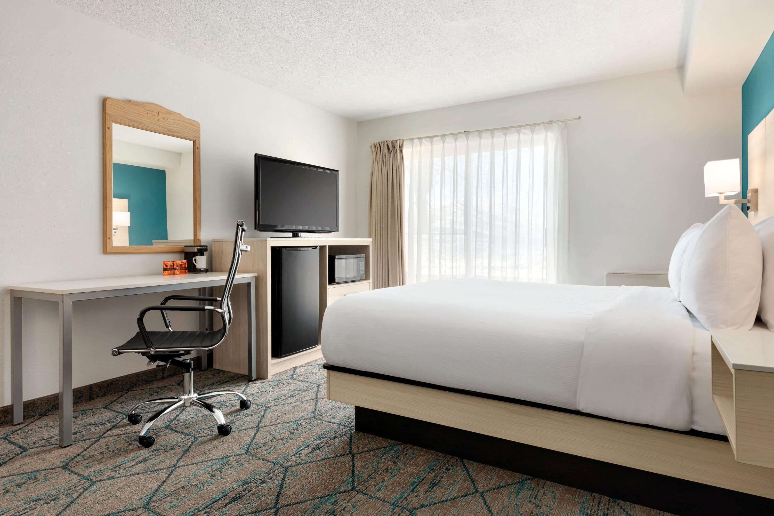 A hotel room with a bed, a desk with a chair, a television, and a large window with curtains.