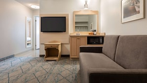 Quarto de hotel com sofá, televisão de tela plana fixada na parede, armário de madeira, micro-ondas e carpete estampado.