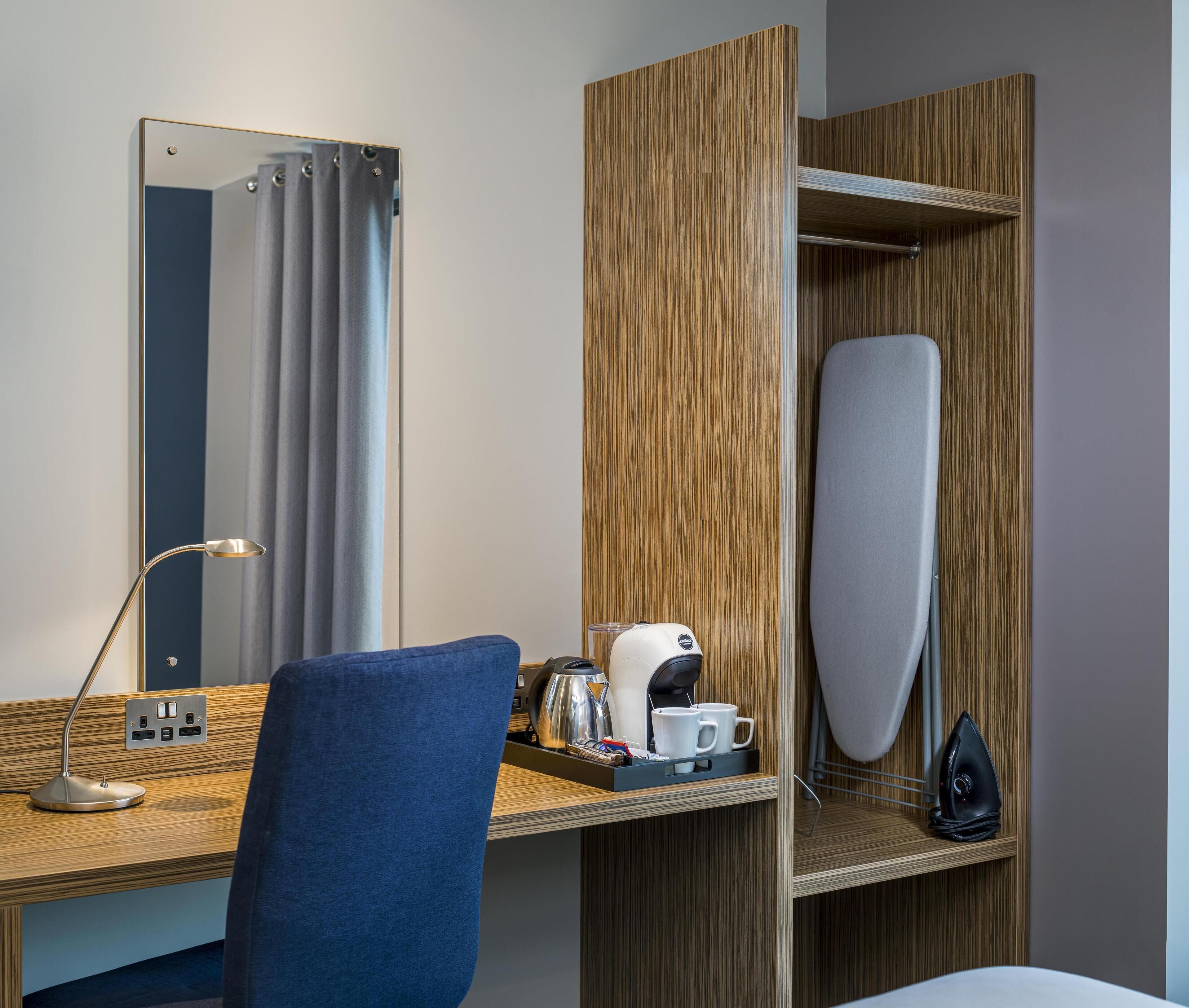 A hotel room with a wooden desk, a mirror, a blue chair, a coffee maker, a kettle, and a hanging iron.