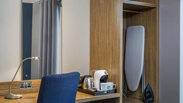 A hotel room with a wooden desk, a mirror, a blue chair, a coffee maker, a kettle, and a hanging iron.