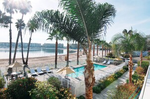 Zona de piscina con sillones de descanso y sombrillas, rodeada de palmeras y vista a la playa y al mar.