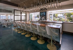 A modern bar area with a long counter, white bar stools, and a television screen above the bar. - Jamaica Bay Inn Marina Del Rey,Tapestry Collection by Hilton (Marina del Rey)