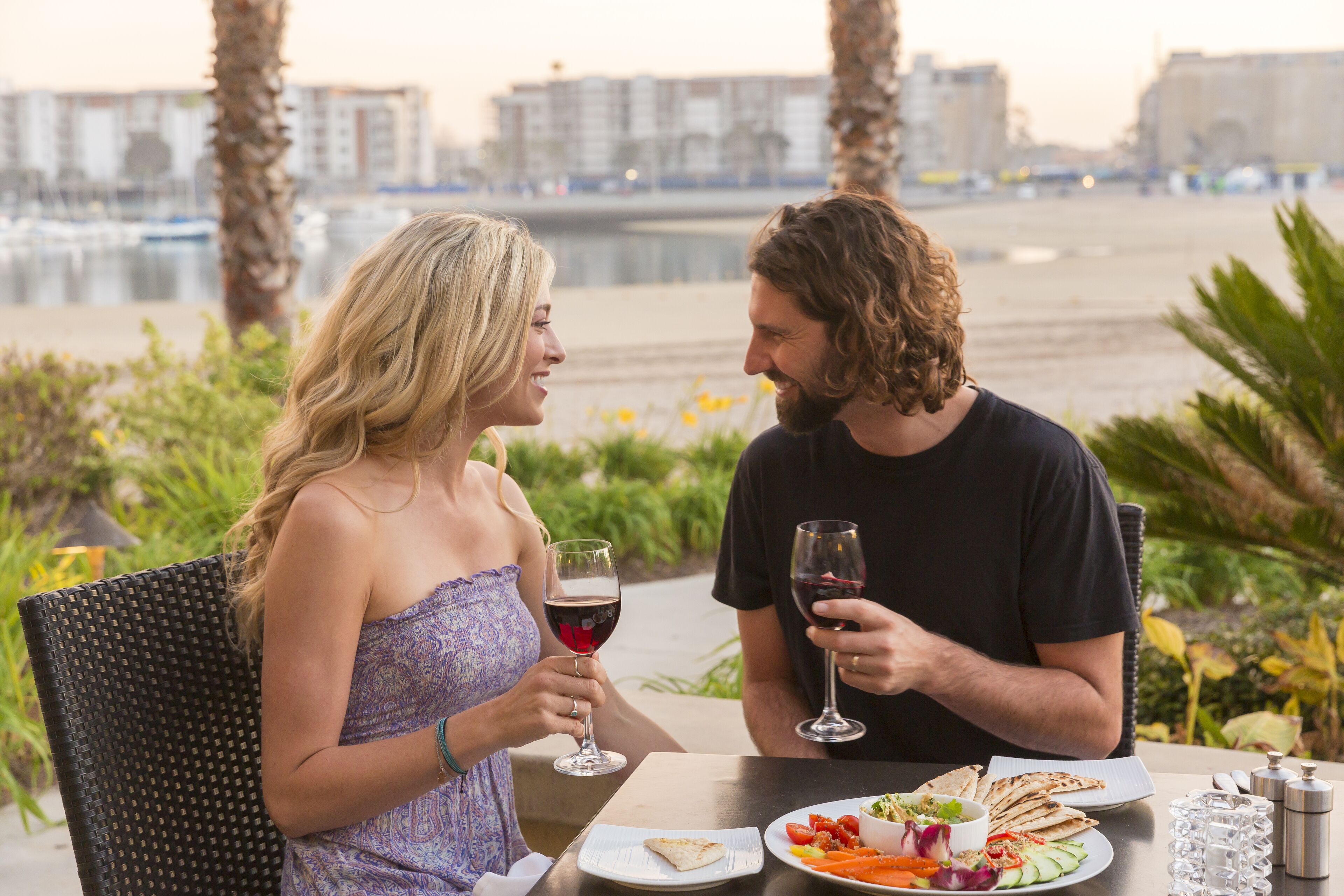 Deux personnes sont assises à une table en plein air, avec vue sur la ville, et tiennent chacune un verre de vin.