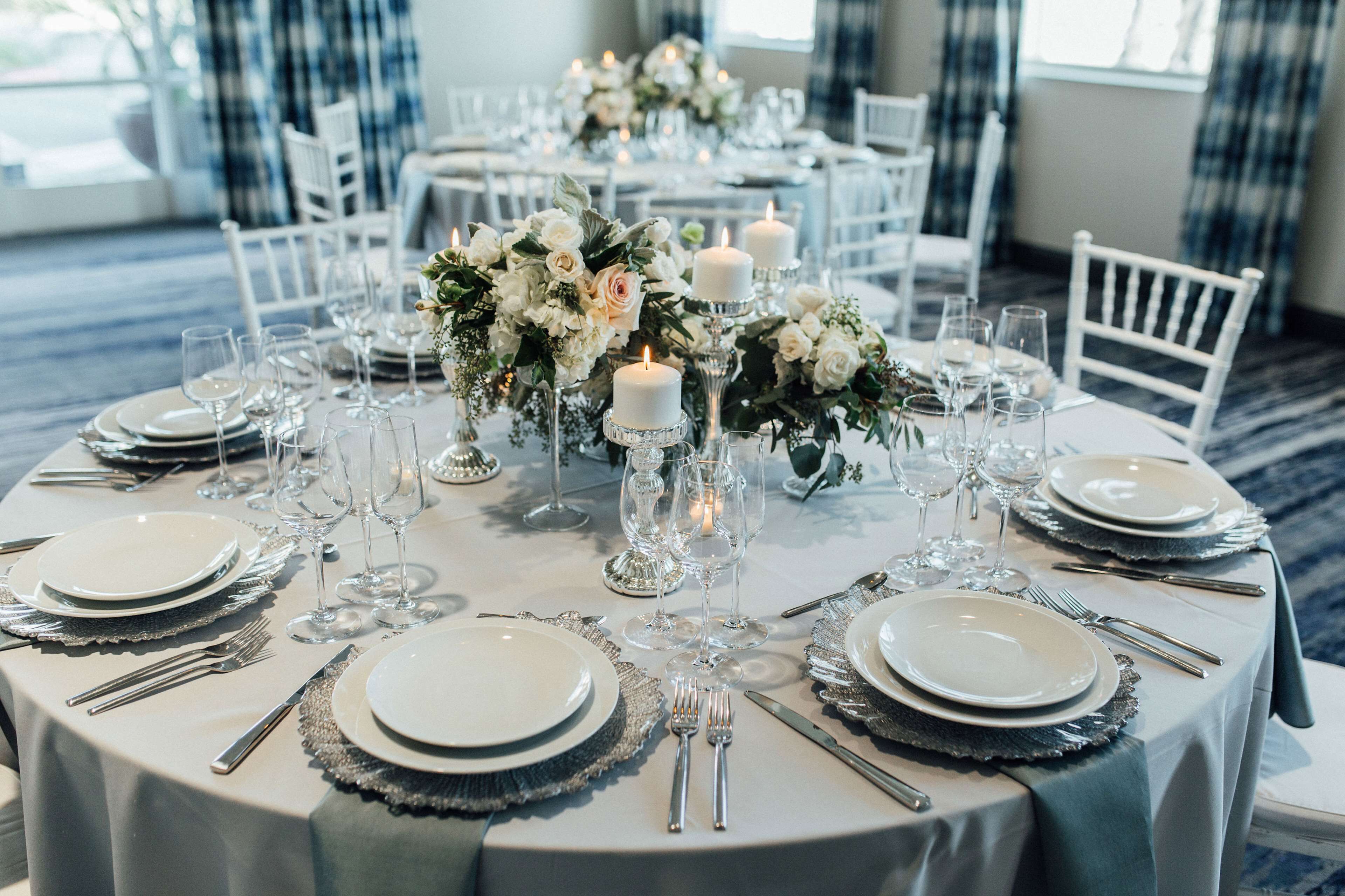 A round dining table set with white plates, silverware, and glassware, surrounded by white chairs.