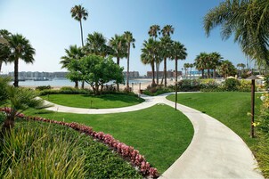 Un jardín ajardinado con un sendero curvo, palmeras y vista a la playa y edificios a lo lejos.