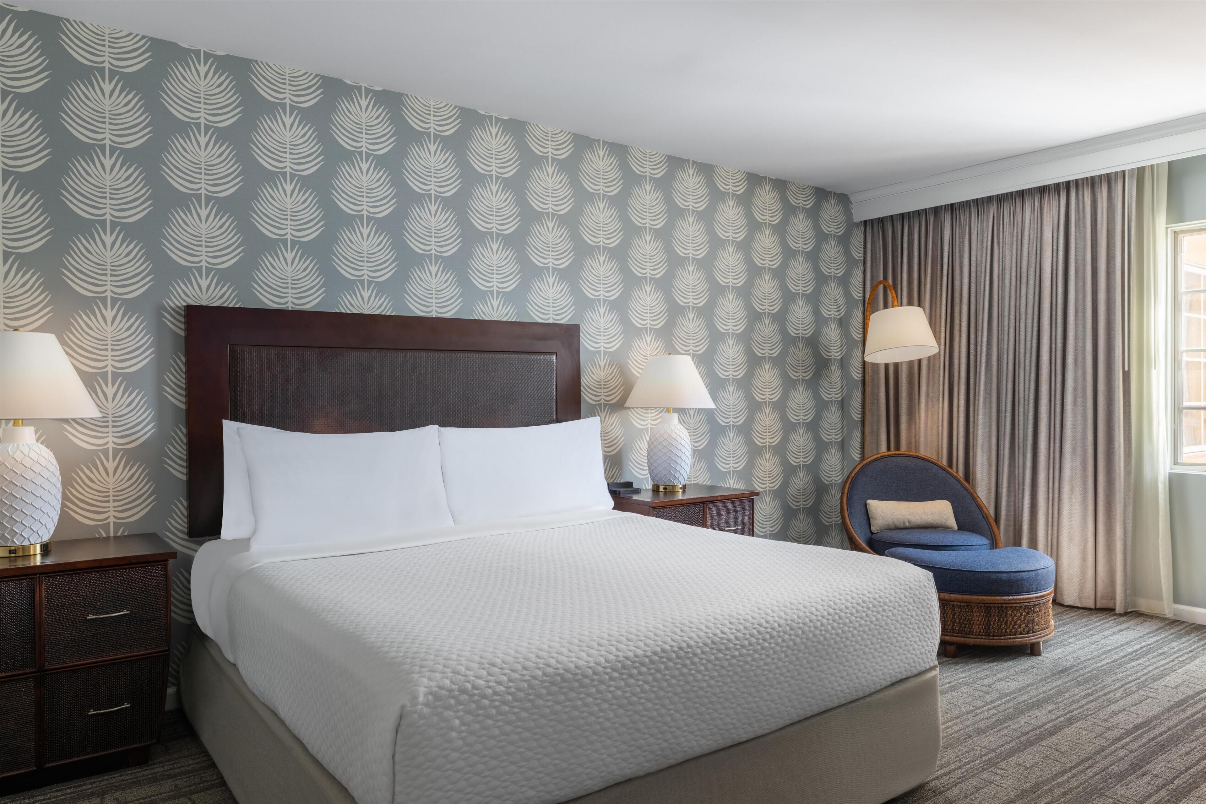 A neatly made bed with white linens, a dark wooden headboard, two bedside lamps, a wicker chair, and a patterned wallpaper.