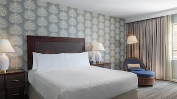 A neatly made bed with white linens, a dark wooden headboard, two bedside lamps, a wicker chair, and a patterned wallpaper.