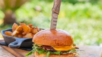 A burger on a wooden board with a knife, a bowl of fries, and a pot on a table with a blurred outdoor background.