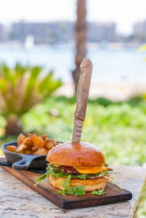 En burger på en træplade med en kniv, en skål pommes frites og en gryde på et bord med en uskarpt udendørs baggrund.