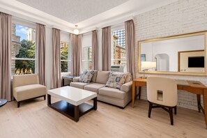 A modern living room with a sofa, armchair, coffee table, and a desk with a chair. Large windows offer a view of the cityscape. - Hotel Union Square (San Francisco)