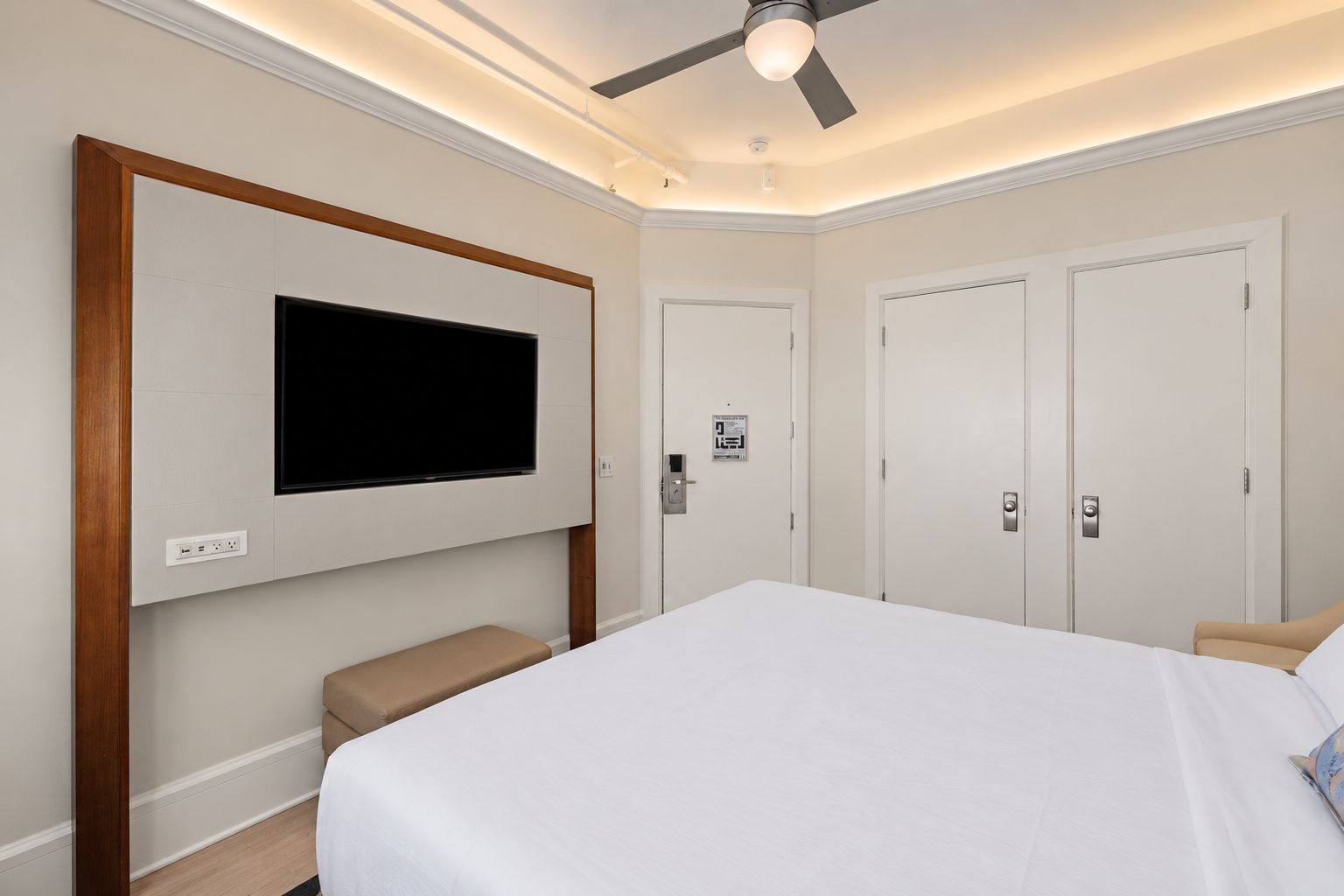 A hotel room with a large bed, a flat-screen TV mounted on the wall, a ceiling fan, and a bench.