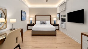 A hotel room with a bed, bedside tables, a desk, a chair, a television, a ceiling fan, and artwork on the walls.