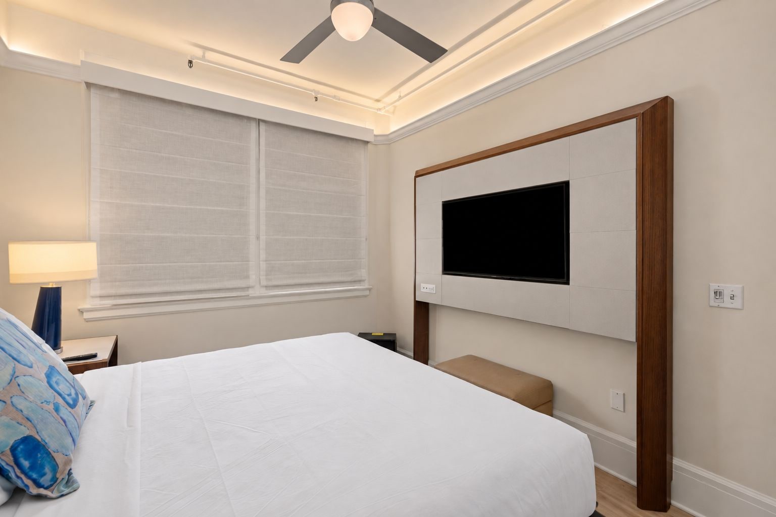 A hotel room with a bed, a TV mounted on the wall, a ceiling fan, and a window with blinds.