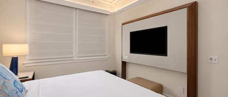 A hotel room with a bed, a TV mounted on the wall, a ceiling fan, and a window with blinds.