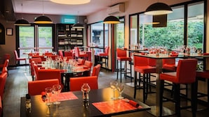 A restaurant interior with red chairs, black tables, and wine glasses. There are large windows with a view of greenery outside.