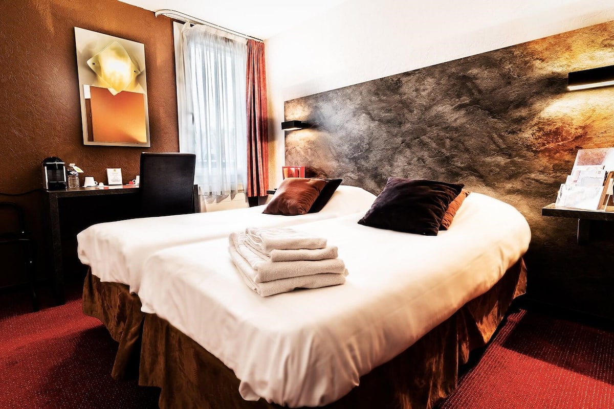 A hotel room with a large bed, a desk with a chair, a coffee machine, and a wall-mounted TV.