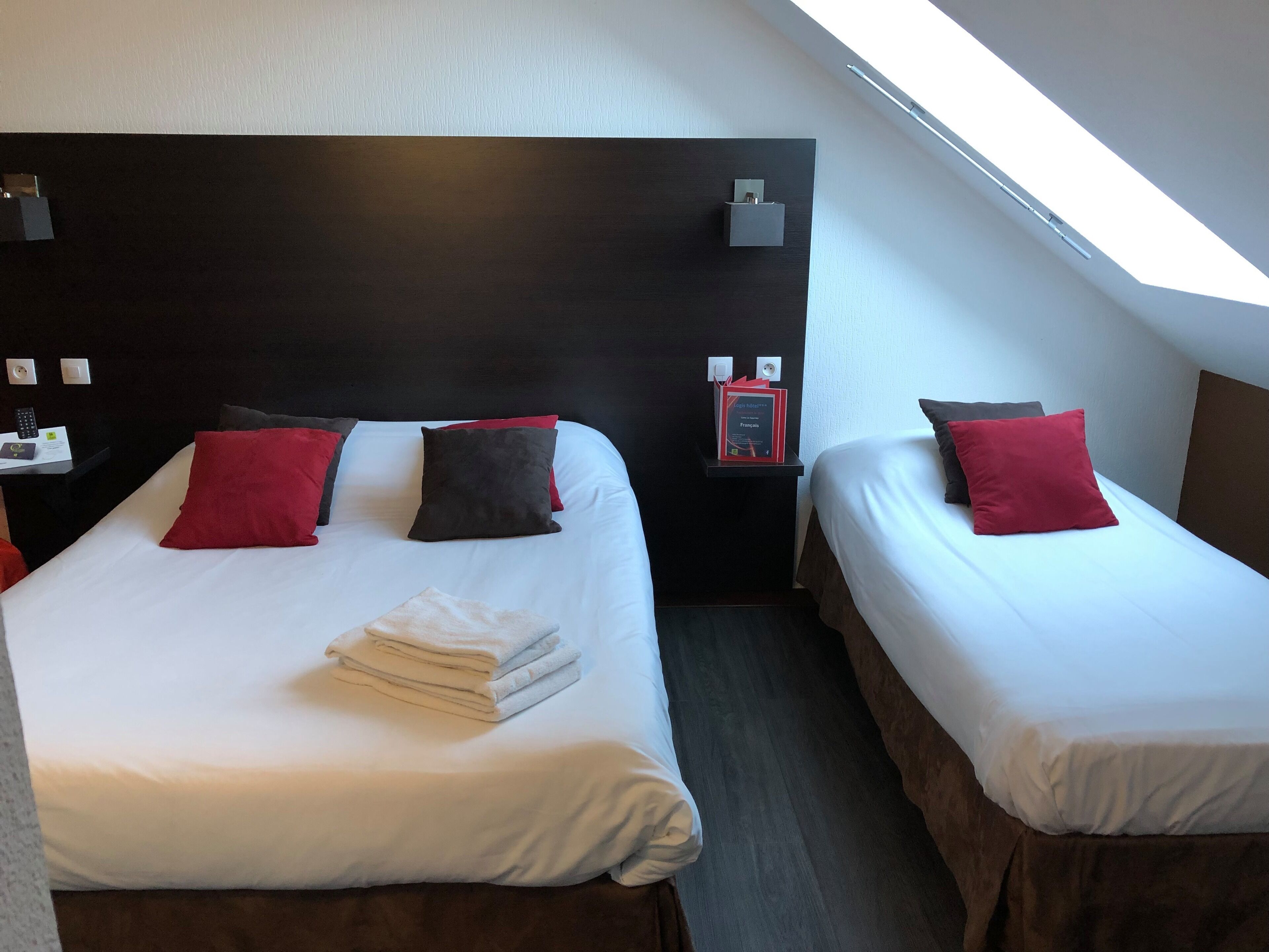 Ein Hotelzimmer mit zwei Betten, jeweils mit weißer Bettwäsche und roten Kissen. Ein kleiner Tisch trägt eine Lampe und ein Buch. Das Zimmer hat eine dunkle Kopfteil und ein Oberlicht.