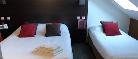 Habitación de hotel con dos camas, cada una con ropa de cama blanca y almohadas rojas. Hay una mesita pequeña con una lámpara y un libro encima. La habitación tiene un cabecero oscuro y un tragaluz.