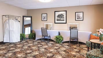 A hotel room with a large mirror, a vanity table with lights, a chair, a sofa, and a patterned carpet.