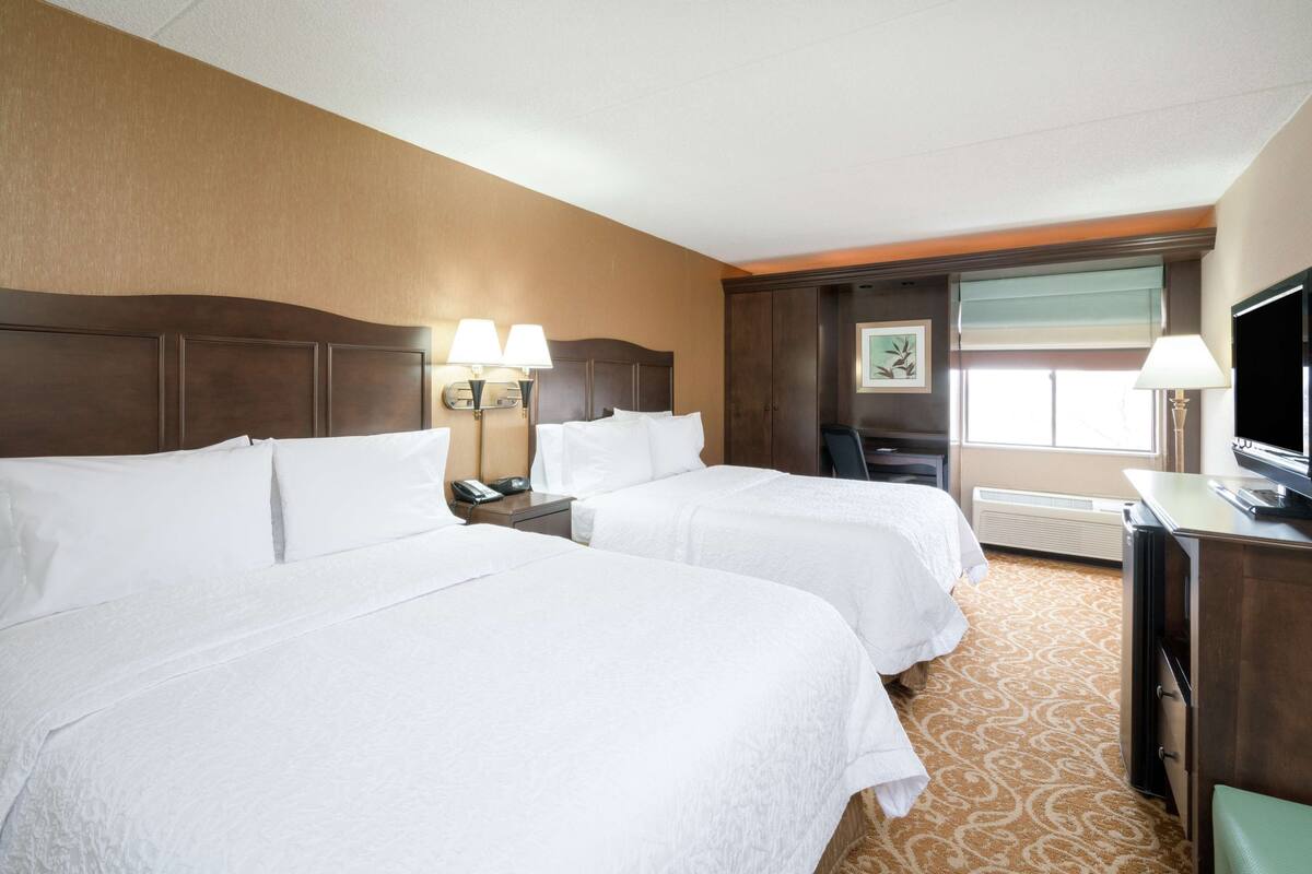 A hotel room with two beds, a desk, a chair, a TV, and a window with blinds.