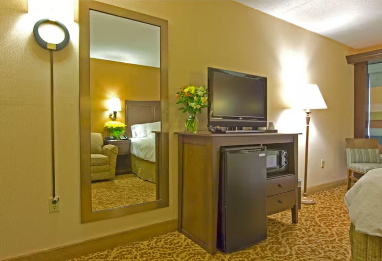 A hotel room with a bed, a desk with a television, a refrigerator, a lamp, and a mirror reflecting the room.