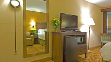 A hotel room with a bed, a desk with a television, a refrigerator, a lamp, and a mirror reflecting the room.