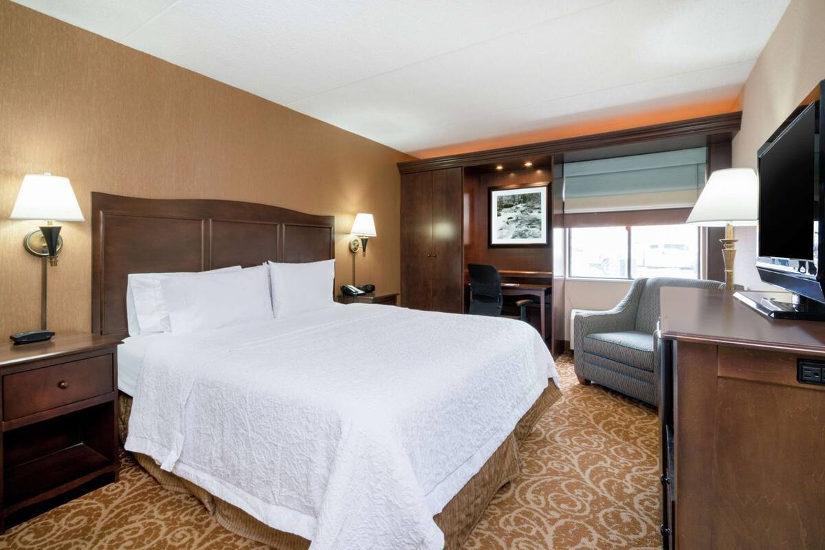 A hotel room with a large bed, two bedside lamps, a desk with a chair, a television, and a window with a view.