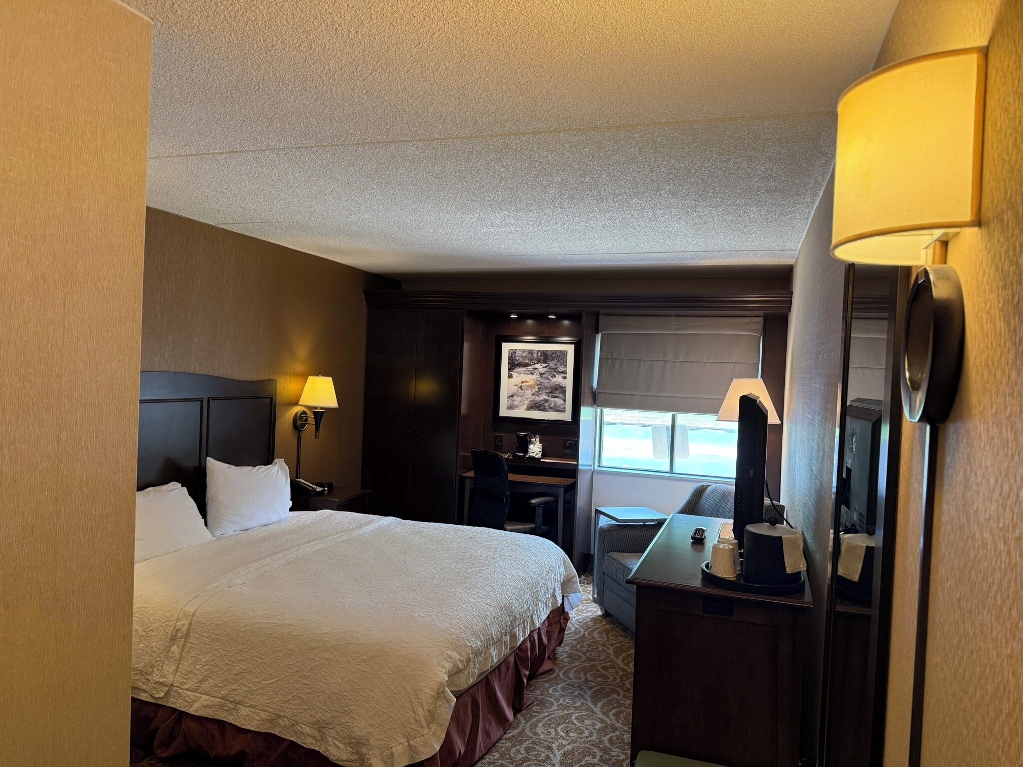 A hotel room with a bed, desk, chair, lamp, and a window with a view.