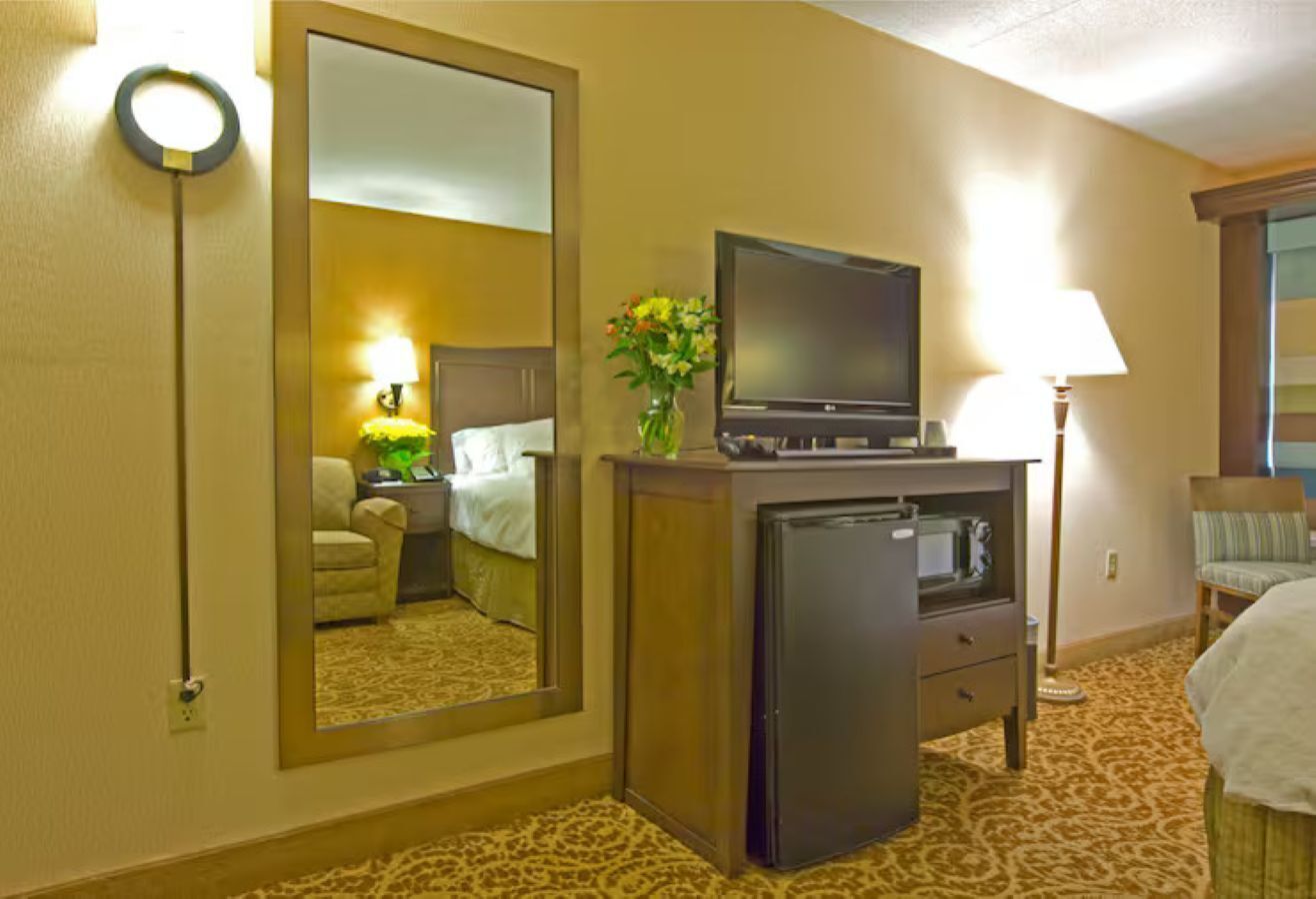 Une chambre d’hôtel comprenant un lit, un bureau avec une télévision, un réfrigérateur, un miroir et une lampe.