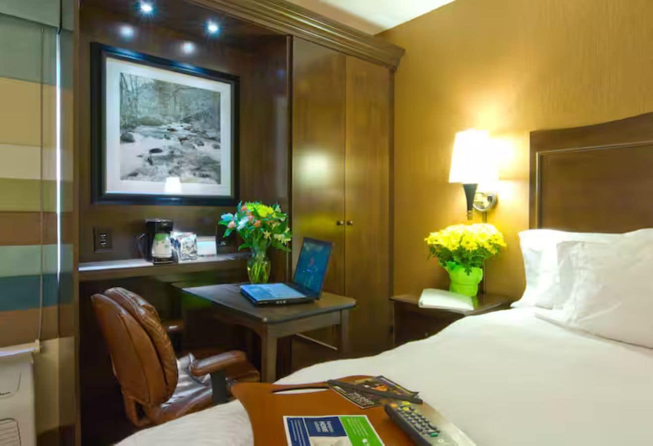 A hotel room with a bed, desk, chair, laptop, and a framed picture on the wall.