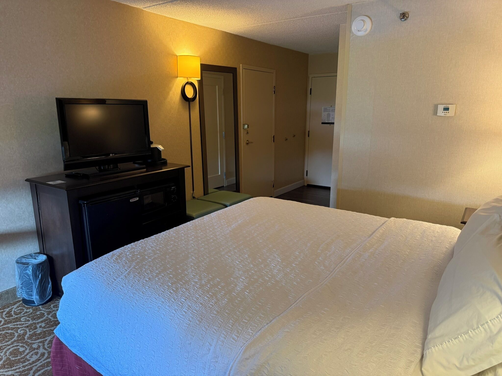 A hotel room with a bed, a television on a stand, a wall-mounted light, and a door.