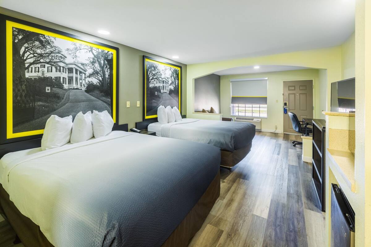 A hotel room with two beds, a desk, a chair, a TV, and large framed pictures on the walls.