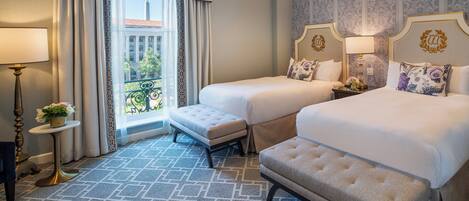 A hotel room with two beds, a small table with a lamp, a bench, and a view of a building through a window.