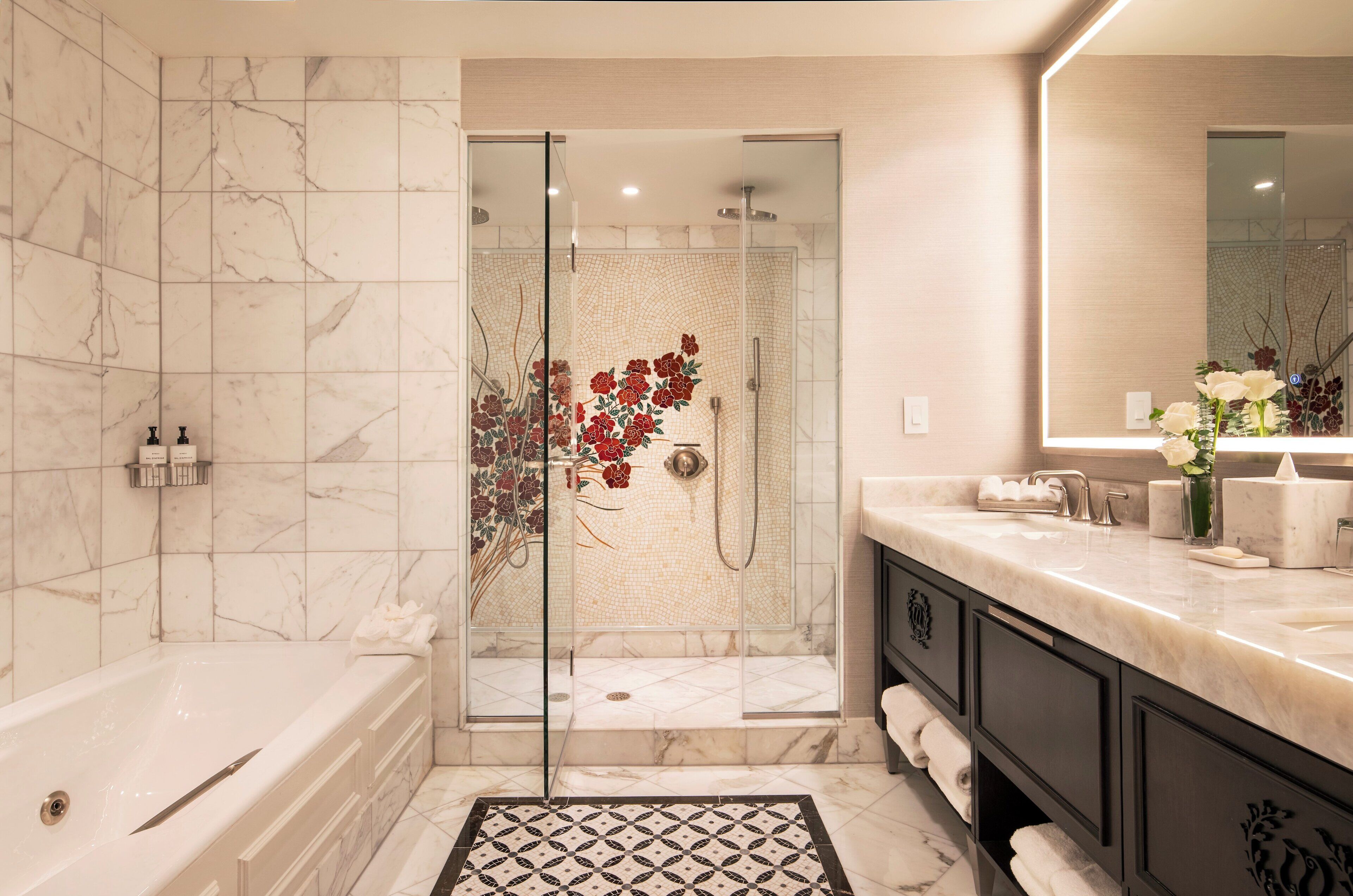 Un baño moderno con ducha envuelta en vidrio, azulejos de mármol, un espejo grande y una encimera de mármol con artículos de aseo.
