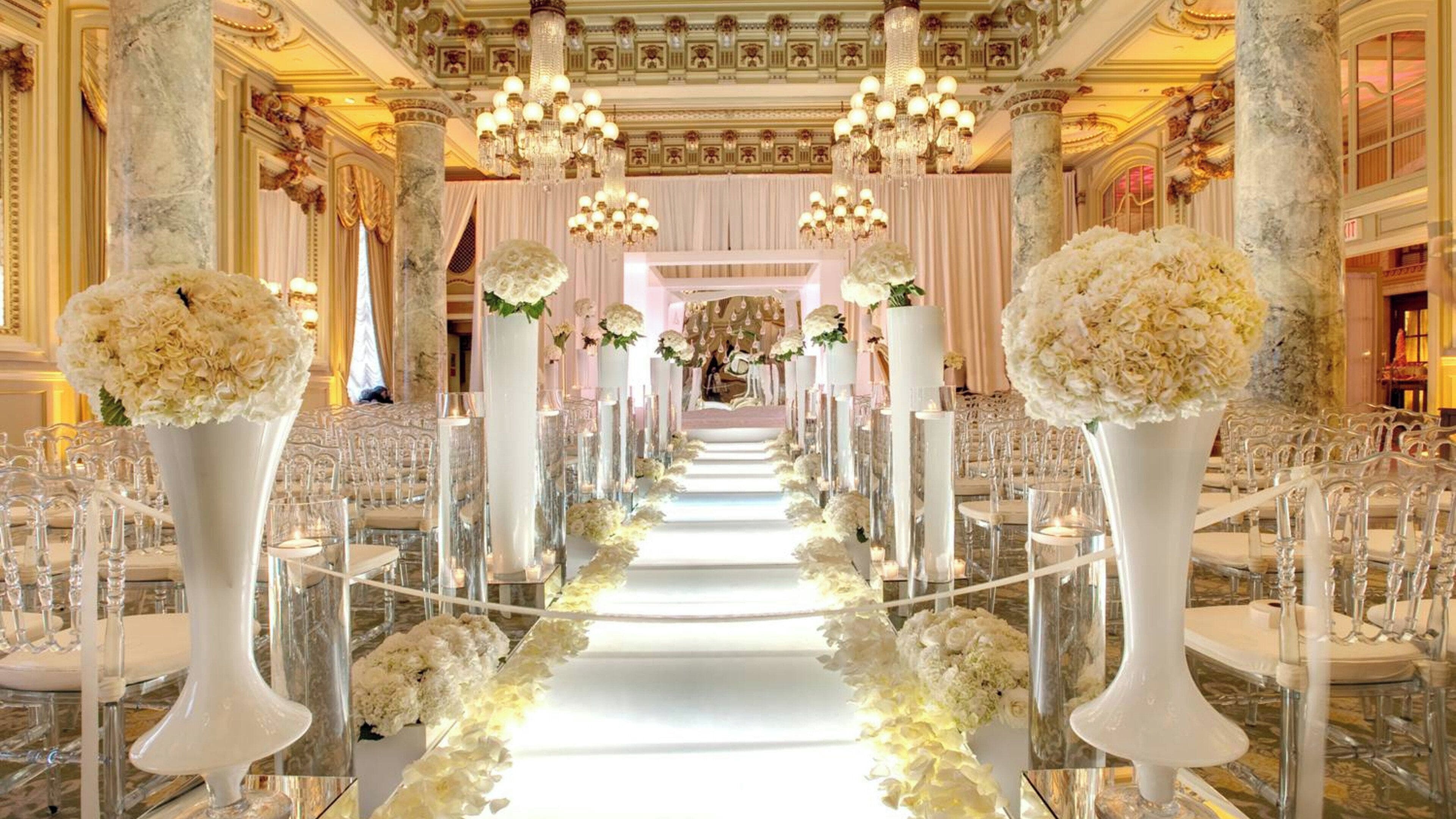 A grand hall with a central aisle, flanked by rows of chairs, and adorned with tall vases of flowers.