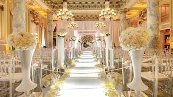 A grand hall with a central aisle, flanked by rows of chairs, and adorned with tall vases of flowers.
