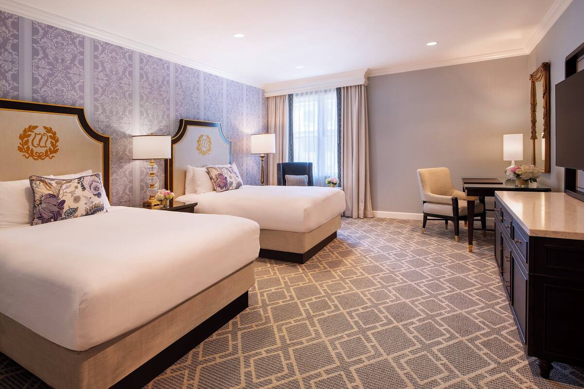 A hotel room with two beds, a desk, a chair, and a TV.