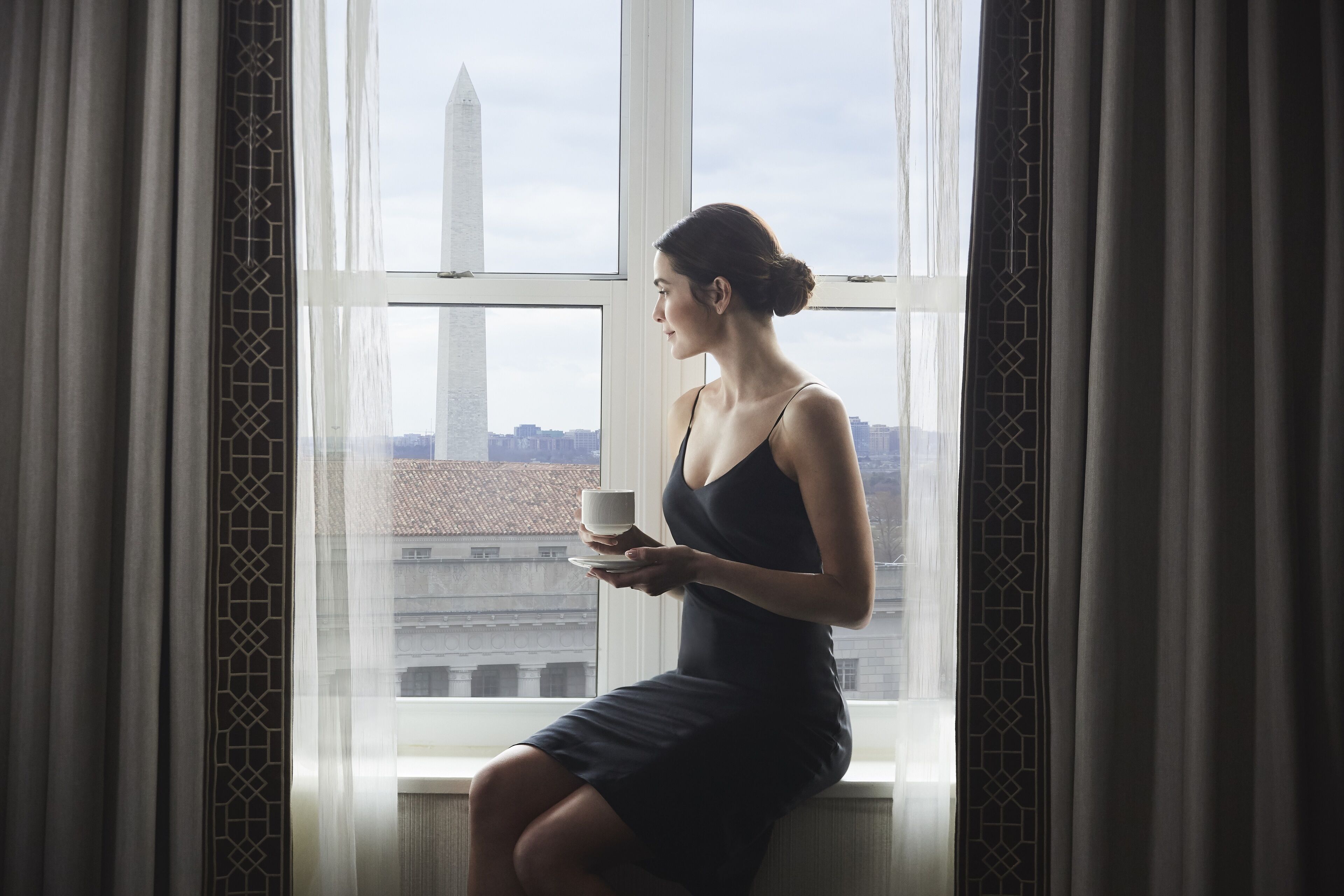 Eine Frau sitzt am Fenster mit Blick auf eine Stadtlandschaft und ein Denkmal.