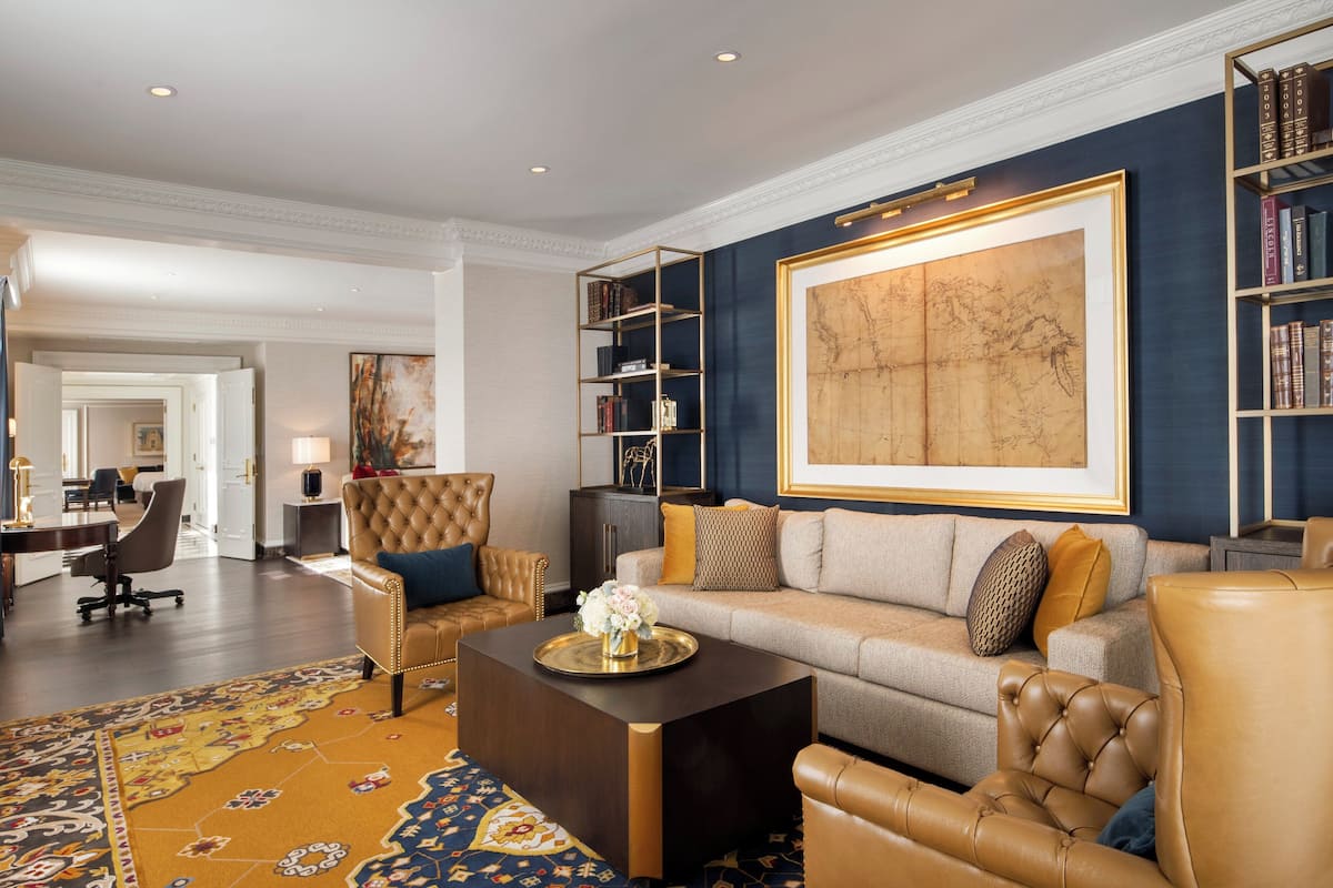 A spacious living room with a large sofa, a coffee table, and a bookshelf. There is a large painting on the wall and a rug on the floor.